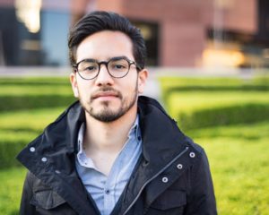 man posing for portrait headshot with jacket and green shrubs and red brick building